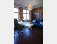 Arbeitszimmer mit Ziegelwand, ausgestattet mit einem weißen Schreibtisch, einem schwarzen Bürostuhl und einer blauen Couch mit Kissen. Ein dekorativer Kronleuchter hängt von einer weiß gestrichenen Decke, und große Fenster lassen Tageslicht in den Raum.