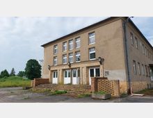 Ein zweigeschossiges Gebäude mit schlichter, beiger Fassade und rechteckigen weißen Fenstern. Der Eingangsbereich hat drei Türen und wird von gemauerten Pfeilern sowie kleinen Treppenstufen flankiert; im Hintergrund sind Bäume und eine Wiese sichtbar.