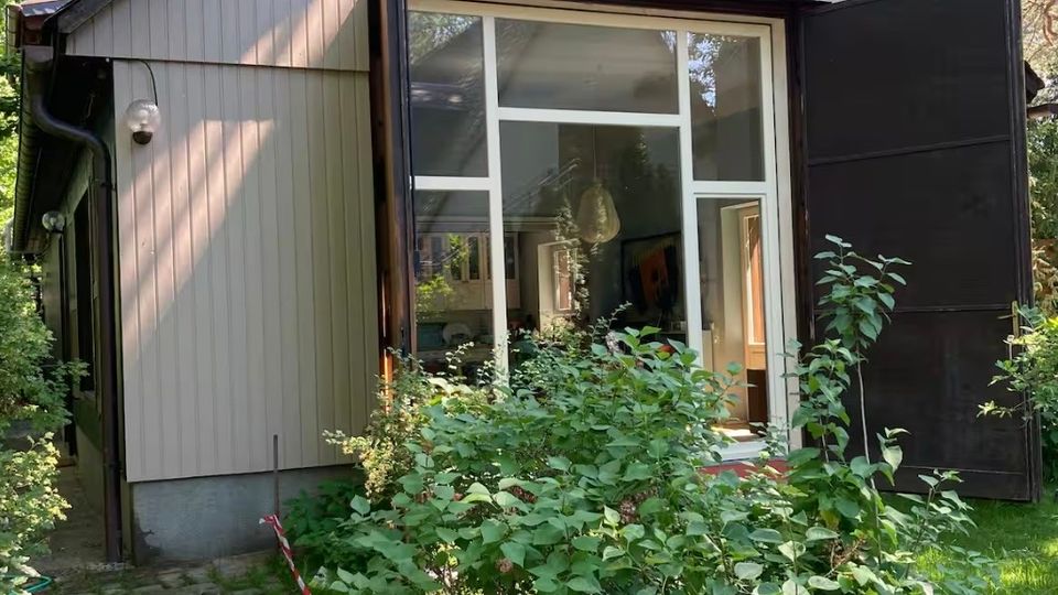 Kleines Holzgebäude mit Satteldach und hellen Fassadenbrettern, dessen große, verglaste Front von zwei aufgeklappten dunklen Türen eingerahmt wird. Davor führen ein gepflasterter Weg und dichter Gartenbewuchs mit Sträuchern und Bäumen durch eine grüne Umgebung.