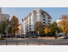 Straßenecke in einem städtischen Wohngebiet mit einem großen, mehrstöckigen Eckgebäude im klassischen Stil, das über weiße Fassaden und rote Dachschindeln verfügt. Umgeben von Bäumen mit herbstlich gefärbtem Laub sind Fahrräder, parkende Autos und Fußwege zu erkennen.