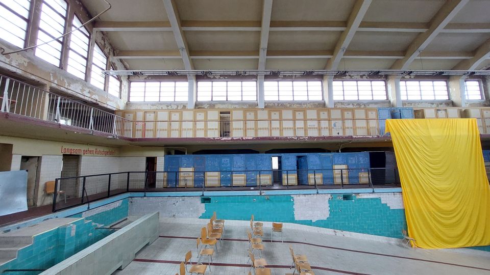 Ein leerstehendes, ehemaliges Hallenbad mit einer leeren Schwimmbeckenmulde, umgeben von verblassten türkisfarbenen Fliesen. Im Hintergrund sind Umkleidekabinen und große Fenster mit Rostspuren an den Wänden, sowie ein gelber Stoffvorhang rechts sichtbar.
