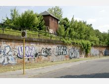 Backsteinmauer mit Graffiti, daneben ein Gehweg und eine Straße; darüber eine grüne Böschung mit Bäumen und Gebüsch, hinter der ein kleines Backsteingebäude mit Walmdach sichtbar ist. Im Vordergrund steht ein Haltestellen-Schild.