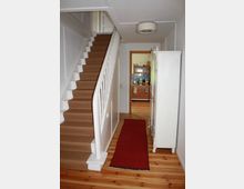 Ein schmaler Hausflur mit Holzboden, einem roten Läufer und weißen Wänden. Links führt eine mit Teppich belegte Treppe nach oben, rechts steht ein weißer Schrank, und im Hintergrund ist eine Türöffnung, die in eine Küche führt.