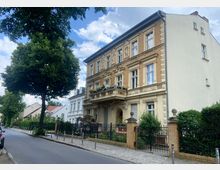 Historisches Wohngebäude mit gelber Fassade und dekorativen Stuckelementen, einem Balkon mit Blumen, sowie einem Vorgarten mit Zaun. Das Haus befindet sich an einer ruhigen, baumgesäumten Straße mit weiteren Wohnhäusern im Hintergrund.