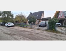 Ein Einfamilienhaus mit orangefarbener Fassade und grauem Satteldach, umgeben von einem Garten mit Bäumen und einem Metallzaun. Vor dem Haus befinden sich zwei Pkw auf gepflasterten Stellplätzen, und die Straße davor ist unbefestigt.