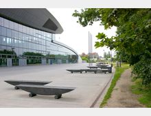 Weitläufiger Vorplatz eines modernen, geschwungenen Gebäudes mit einer Fassade aus Glas und Metall, umgeben von Bäumen und Grünflächen; im Vordergrund befinden sich skulpturale Sitzbänke und Metallstangen.