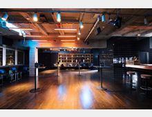 Moderner Veranstaltungsraum mit Holzboden, vereinzelten Stehtischen und einer Bar mit schwarzen Fliesen an der rechten Wand. Im Hintergrund befindet sich ein Regal mit Deko-Elementen und Sitzmöglichkeiten, über dem Raum sind Beleuchtungselemente an einer sichtbaren Betondecke angebracht.