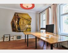 Ein helles Arbeitszimmer mit Holzdielenboden, einem großen Schreibtisch aus Holz mit schwarzem Gestell, auf dem ein Computer und eine Schreibtischlampe stehen. An der Wand hängt ein modernes Kunstwerk, darunter stehen gerahmte Bilder, und die Fenster sind mit hellen Vorhängen versehen.