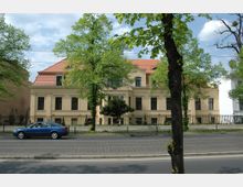 Historisches Gebäude mit heller Fassade und rotem Ziegeldach, umgeben von einem Zaun und Bäumen entlang einer Straße. Im Vordergrund fährt ein blauer Pkw über den Asphalt, während der Himmel leicht bewölkt ist.