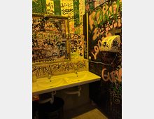 Kleiner Sanitärraum mit Doppelwaschbecken und großem Spiegel, dessen Rahmen und Glas mit zahlreichen Graffiti und Aufklebern bedeckt sind. Die Wände sind vollständig mit bunten Tags und Schriftzügen überzogen; rechts hängt ein Wandspender neben sichtbaren Leitungen.