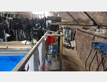 Ein Innenraum mit Taucherausrüstung wie Tarierwesten und Flossen, die an Metallständern aufgehängt sind, und einer Arbeitsplattform aus Metall mit Geländern neben einem Wasserbecken. Die Wände bestehen aus sichtbaren Ziegelsteinen, und an der Seite befinden sich Schläuche und Schilder.