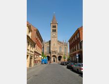 Straßenansicht einer historischen Kirche mit Backsteinfassade und einem hohen Glockenturm mit Turmkreuz. Links und rechts flankieren mehrstöckige Gebäude mit roten und gelben Fassaden die gepflasterte Straße, auf der Autos parken.