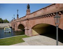 Rote Sandsteinbrücke mit verzierten Bögen und Steinfiguren, darunter Laternen entlang des Geländers; im Vordergrund ein gepflasterter Gehweg und eine Grünfläche neben einem Fluss.
