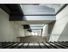 Blick nach unten in ein modernes Treppenhaus mit Betonwänden, offenen Stufen aus Metall und großzügigen Glasflächen, die Tageslicht hereinlassen. Die Struktur zeigt klare Linien und eine industrielle Ästhetik.