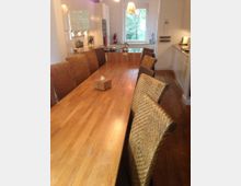 Langer Tisch aus Holz mit geflochtenen Stühlen in einem offenen Küchen- und Essbereich. Im Hintergrund befinden sich helle Küchenschränke, Fenster mit Blick ins Grüne und eine Küchenzeile mit Herd und Spüle.