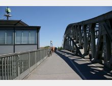 Eine Stahlbrücke mit sichtbaren Trägern und Straßenlaternen führt über eine Fahrbahn und einen Gehweg. Links des Gehwegs befindet sich ein moderner, verglaster Bahnhof mit einem "S-Bahn"-Symbol auf dem Dach, während Fahrräder entlang des Geländers abgestellt sind.