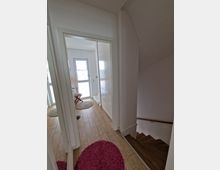 Ein hell beleuchteter Eingangsbereich mit hellbraunem Fliesenboden und weißen Wänden. Rechts führt eine Holztreppe nach unten, während vorne eine Tür mit Glaselementen und ein Spiegel an der Wand sichtbar sind.