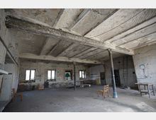 Großer, leerer Industriehallenraum mit freiliegenden, verwitterten Deckenbalken, abblätterndem Putz und mehreren Stahlstützen. Entlang der Backsteinwände befinden sich mehrere Fenster sowie vereinzelt abgestellte Möbel und eine Leiter auf dem Betonboden.