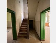 Ein verlassener Hausflur mit abgenutzten Wänden und einer schmalen Holztreppe, die nach oben führt. Links und rechts befinden sich Türöffnungen mit grünen Rahmen, die in leere Räume führen, während am Boden Schmutz und Verfall sichtbar sind.