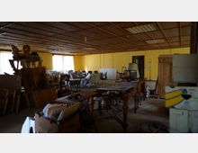 Ein großer Innenraum mit gelb gestrichenen Wänden und einer Holzdecke, gefüllt mit unterschiedlichen Möbelstücken wie Stühlen, Tischen, Kommoden und verpackten Polstermöbeln. Der Raum wirkt wie ein Lager oder eine Abstellfläche, mit Kartons und lose verstauten Gegenständen.