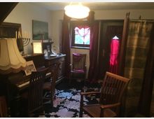 Ein kleiner, gemütlicher Raum mit antiken Möbeln, darunter ein Klavier mit Stehlampe, Holztische und Stühle sowie ein dekorativer Teppich mit Blumenmuster. Vorhänge in Weinrot und eine Trennwand verleihen dem Raum eine private Atmosphäre, während ein Fenster und eine Tür im Hintergrund für Licht sorgen.