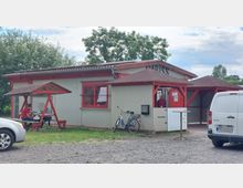 Ein kleiner Imbissstand mit grauen Wänden und roten Akzenten, umgeben von einer ländlichen Umgebung. Vor dem Gebäude steht ein überdachter Sitzbereich mit Tischen, ein abgestelltes Fahrrad und ein weißer Transporter in einer geschotterten Parkfläche.