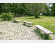 Drei einfache Steinbänke stehen auf einem gepflasterten Platz in einem Park, umgeben von Rasenflächen, Sträuchern und einer Baumallee im Hintergrund. Ein schmaler Fußweg führt durch das grüne Gelände.