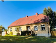 Ein einstöckiges Haus mit gelber Fassade und rotem Satteldach, umgeben von einem großen Garten mit Rasen und Bäumen. Links vom Gebäude befindet sich eine helle Gartentür, und der Himmel ist klar und blau.