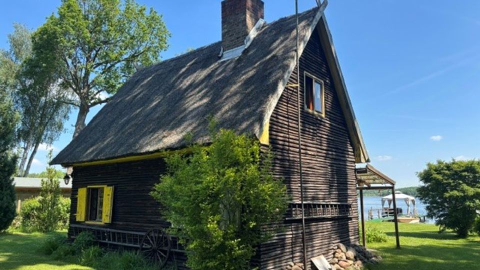 Holzhaus mit dunkler Fassadenverkleidung und steilem, strohgedecktem Satteldach mit Schornstein auf einer grünen Wiese. Umgeben von Bäumen und Sträuchern, mit Blick auf ein Gewässer im Hintergrund.