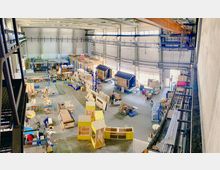 Innenraum einer großen Werkstatt oder Produktionshalle mit hohen Wänden und Oberlichtern. Verschiedene Arbeitsstationen mit Holzwerkstoffen, unfertigen Strukturen, Werkzeugen und Maschinen sind auf dem Boden verteilt.