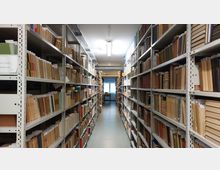 Ein langer Gang in einem Archiv oder einer Bibliothek mit hohen weißen Metallregalen, die beidseitig mit Büchern und Aktenordnern gefüllt sind. Am Ende des Gangs befindet sich ein kleines Fenster, durch das Tageslicht einfällt.