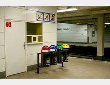 Eine U-Bahn-Station mit einer weißen, gefliesten Wand, auf der Schilder mit Verboten für Rauchen, Radfahren und Kinderwagen angebracht sind. Im Vordergrund stehen vier farbcodierte Mülleimer für Mülltrennung (Restmüll, Papier, Glas, Verpackungen), daneben ein geschlossener Türbereich mit kleinem Fenster.