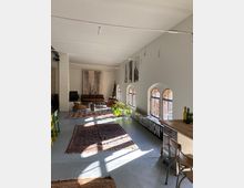 Großer, lichtdurchfluteter Raum mit weiß gestrichenen Wänden und mehreren Fenstern mit Rundbögen, durch die Sonnenlicht einfällt. Der Raum ist mit Teppichen, Holzmöbeln, Pflanzen und Dekoration (darunter ein "Music Television"-Schild) ausgestattet, was eine wohnliche und kreative Atmosphäre schafft.