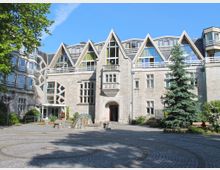 Steingebäude mit moderner Architektur, gekennzeichnet durch spitze Giebel, große Fensterflächen und geometrische Formen. Im Vordergrund ein gepflasterter Platz mit einem kleinen Brunnen sowie Bäumen und Sitzbänken.