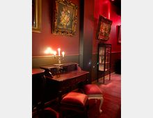 Ein Raum mit klassischer Einrichtung, bestehend aus einem Holzschreibtisch mit einer brennenden Kerze auf einem Leuchter, zwei roten gepolsterten Hockern und dekorativen Gemälden in vergoldeten Rahmen an den dunkelroten Wänden. Im Hintergrund steht ein Glasschrank mit ausgestellten Gegenständen.