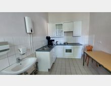 Kleine Küche mit weißen Schränken und einer Arbeitsplatte, ausgestattet mit Spüle, Herd, Kaffeemaschine und Geschirr. Im Vordergrund ein Waschbecken, an der Wand ein Durchlauferhitzer; im rechten Bereich stehen ein Tisch und ein Stuhl.