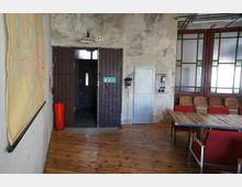 Ein rustikaler Raum mit Holzboden, einer Tür aus Metall mit einem Notausgangsschild darüber und einer Betonwand, an der ein altes Telefon und Schaltanlagen montiert sind. Rechts befinden sich eine Glaswand mit rot-grünen Rahmen und mehrere Holzbänke, links ist ein großes Wandposter sichtbar.