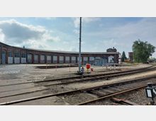 Ein historisches Eisenbahndepot mit einer halbkreisförmigen roten Backsteinhalle, deren Tore geschlossen sind. Im Vordergrund befinden sich Gleise und ein Bahnhofsbereich mit Schildern und Verkehrsabsperrungen, im Hintergrund sind ein alter Wasserturm und Bäume sichtbar.