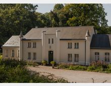 Helles, zweigeschossiges Landhaus mit steilem Schieferdach, symmetrischer Fassade und zentralem Eingang, flankiert von zwei zylindrischen Türmchen und kleinen Eckspitzen. Davor liegt eine Kiesauffahrt mit niedrigen Beeten und Sträuchern, im Hintergrund dichter Baumbestand.