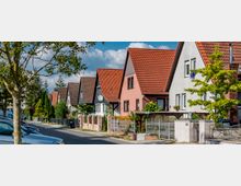 Straßenansicht einer Reihenhaussiedlung mit freistehenden Einfamilienhäusern im Stil der Nachkriegszeit. Die Häuser haben steile Satteldächer mit roten Dachziegeln, Vorgärten mit Zäunen, und parkende Autos säumen die Straße unter vereinzelten Bäumen.