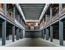 Großer, dunkler Innenraum eines industriellen Veranstaltungsraums mit hoher Decke, sichtbarer Stahlkonstruktion, mehreren Ebenen mit Metallgeländern und einer Bühne im Hintergrund. Die Wände sind schwarz gestrichen, das Licht ist gedämpft und strukturelle Träger verlaufen entlang der Seiten.