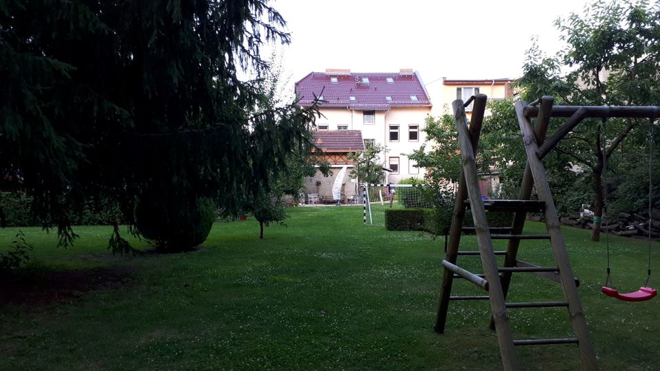 Großer, grasbewachsener Gartenhof mit hohen Bäumen und einem Holzspielgerät mit Schaukel im Vordergrund. Im Hintergrund stehen mehrstöckige Wohnhäuser mit hellen Fassaden und roten Ziegeldächern sowie ein kleiner Fußballrahmen auf der Wiese.