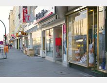 Straßenszene mit einer Reihe von Geschäften, darunter ein Telekom-Laden, eine McDonald's-Filiale und weitere Einzelhandelsgeschäfte. Schaufenster zeigen ausgestellte Waren, wie Kleidung und Unterwäsche, und der Bürgersteig ist mit Passanten, Müllcontainern und Verkehrsschildern belebt.