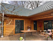 Blockhütte mit Holzfassade und grünem Metalldach, umgeben von kahlen Ästen. Der Außenbereich zeigt eine Terrasse mit Holzboden, einem Tisch, einer Sitzecke mit Polstermöbeln und dekorativen Pflanzen.