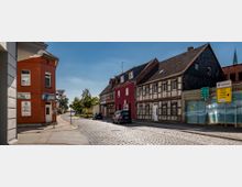 Straßenszene in einer Kleinstadt mit Kopfsteinpflaster, links ein rotes Gebäude mit einem Hotel- und Restaurant-Schild, rechts historische Fachwerkhäuser und ein gemaltes Wandbild. Im Hintergrund sind Bäume und mehrere Wegweiser mit Zielbeschreibungen sichtbar.