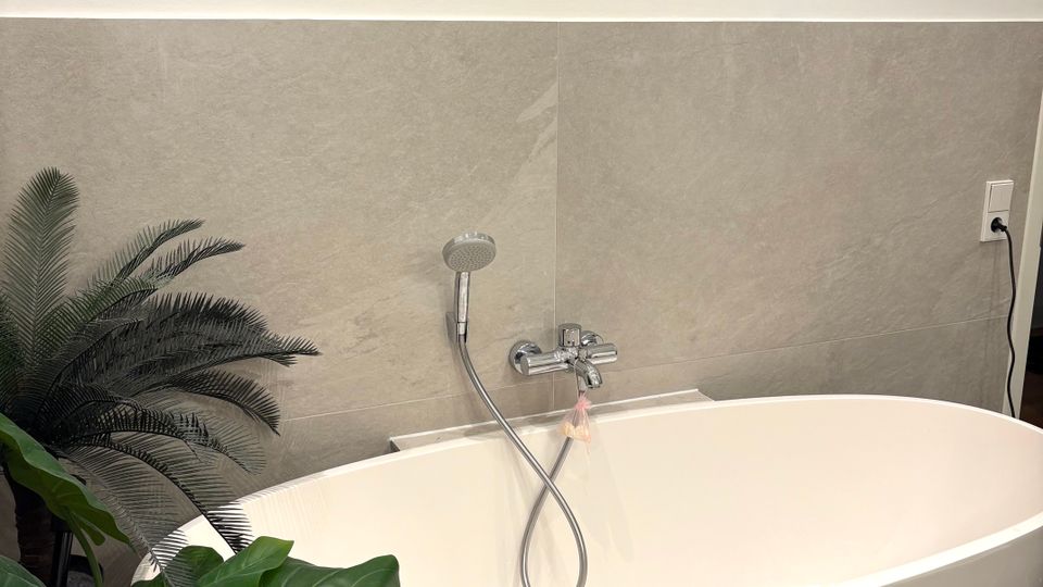 Freistehende weiße Badewanne in einem modernen Badezimmer mit grauen Wand- und Bodenfliesen sowie Wandarmatur mit Handbrause. Links stehen mehrere große Zimmerpflanzen neben der Wanne.