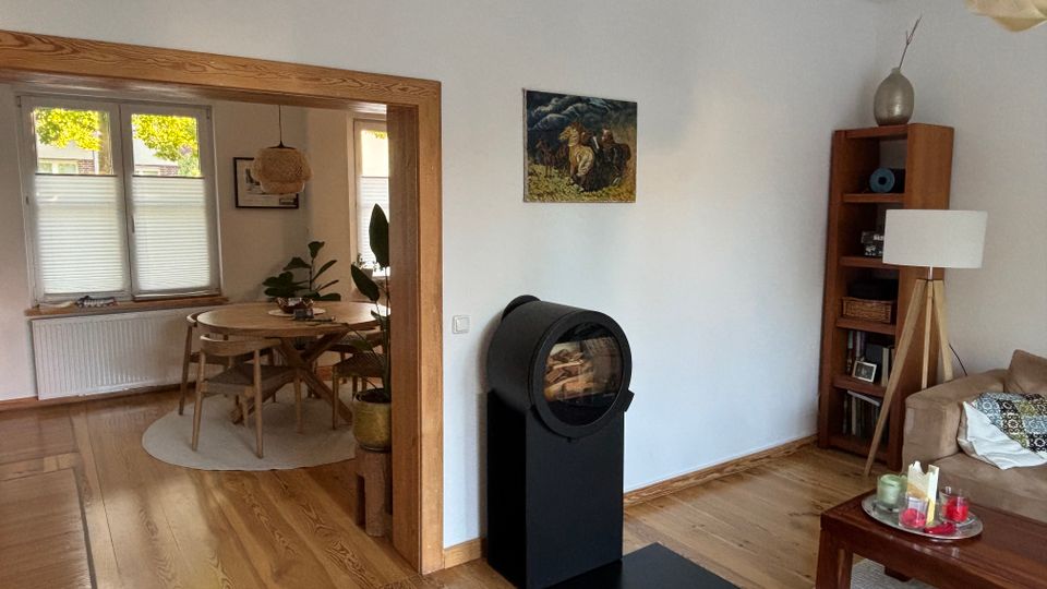 Blick in ein helles Wohn- und Esszimmer mit Holzfußboden: rechts ein schwarzer, runder Kaminofen vor weißer Wand neben Regal, Stehlampe und Sofa. Durch einen breiten Holzrahmen führt der Durchgang in den Essbereich mit rundem Holztisch, Stühlen, Pflanzen und großen Fenstern mit halb geschlossenen Plissees.