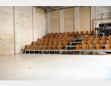 Eine Halle mit sichtbaren weißen Ziegelwänden und einer erhöhten Sitztribüne aus Holzstühlen auf Metallgestellen. Der Raum hat einen neutralen, hellen Boden und eine industrielle Atmosphäre mit Rohren und Technik im Hintergrund.