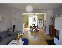 Wohnzimmer mit Blick auf einen Essbereich in einem offenen Wohnkonzept. Der Raum verfügt über einen hölzernen Fischgrätboden, eine graue Couch, eine große weiße Deckenlampe, sowie eine Fensterfront im Hintergrund, die viel Tageslicht hereinlässt; links ein Küchenschrank und rechts ein weißer Kamin sowie ein Sideboard.