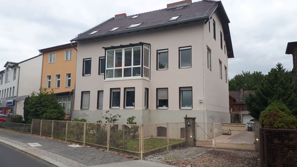 Mehrstöckiges Wohnhaus mit hell verputzter Fassade, Satteldach mit Dachfenstern und einem verglasten Erker an der Straßenseite. Vor dem Gebäude befinden sich ein kleiner, eingezäunter Vorgarten sowie eine Einfahrt mit Tor und gepflastertem Bereich entlang eines Gehwegs.
